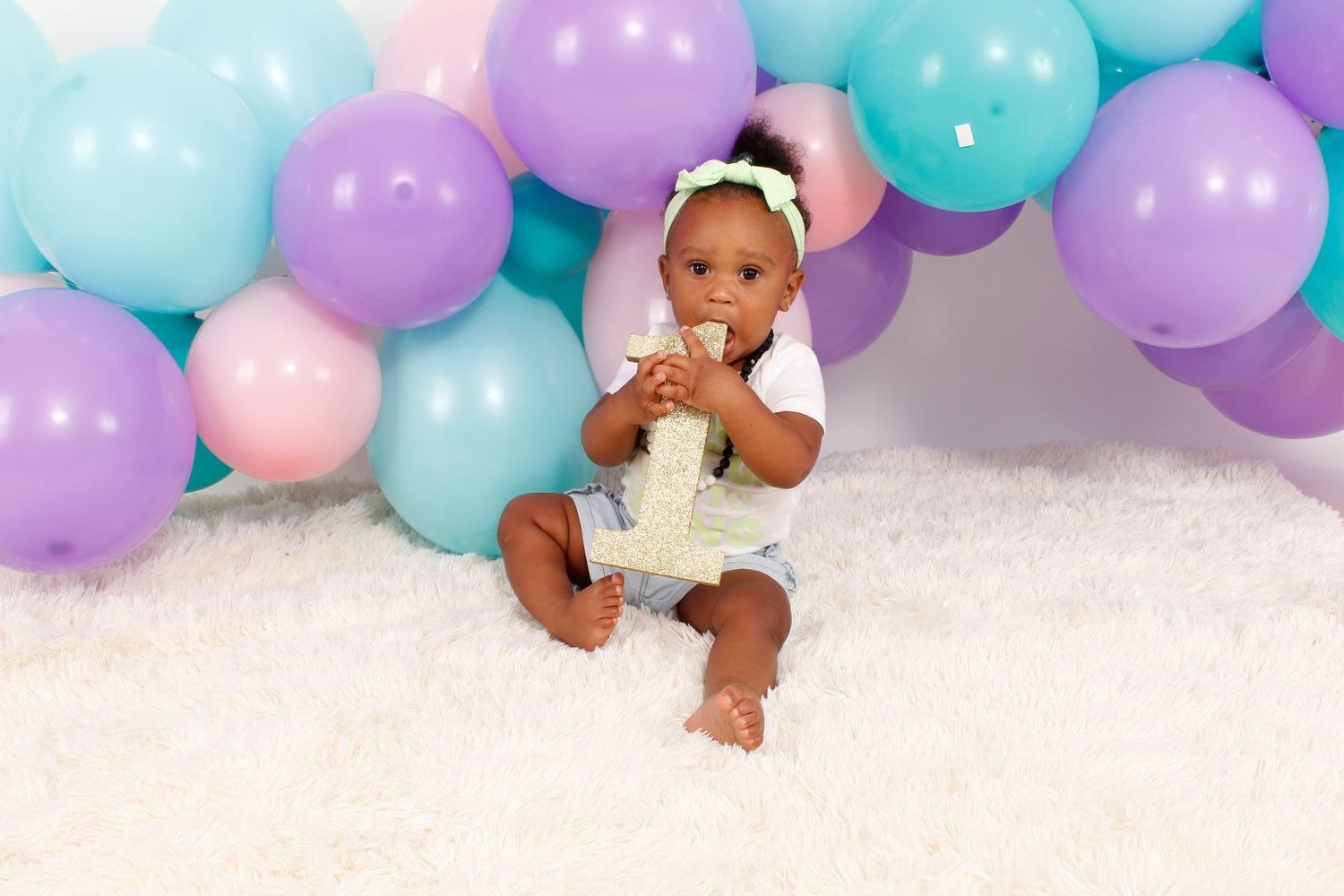 A baby sits on a white rug surrounded by colorful balloons at a birthday celebration.
