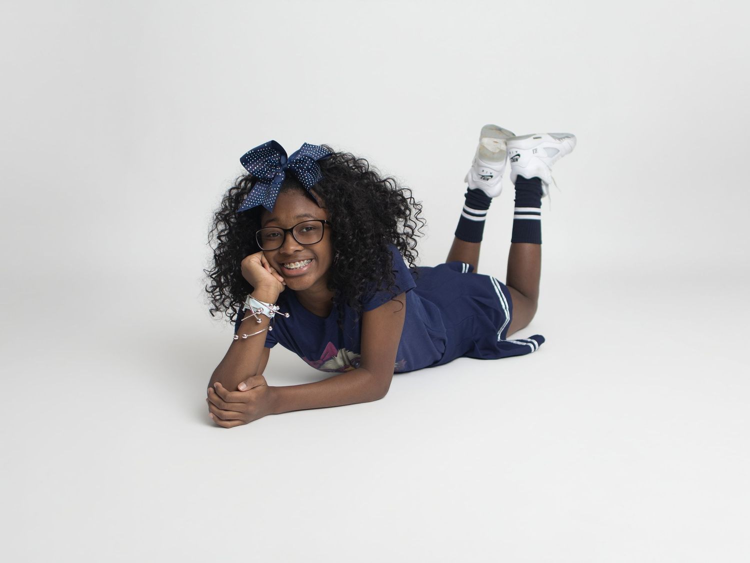 Young girl in navy blue outfit with bow headband, glasses, lying on floor smiling in studio photo.