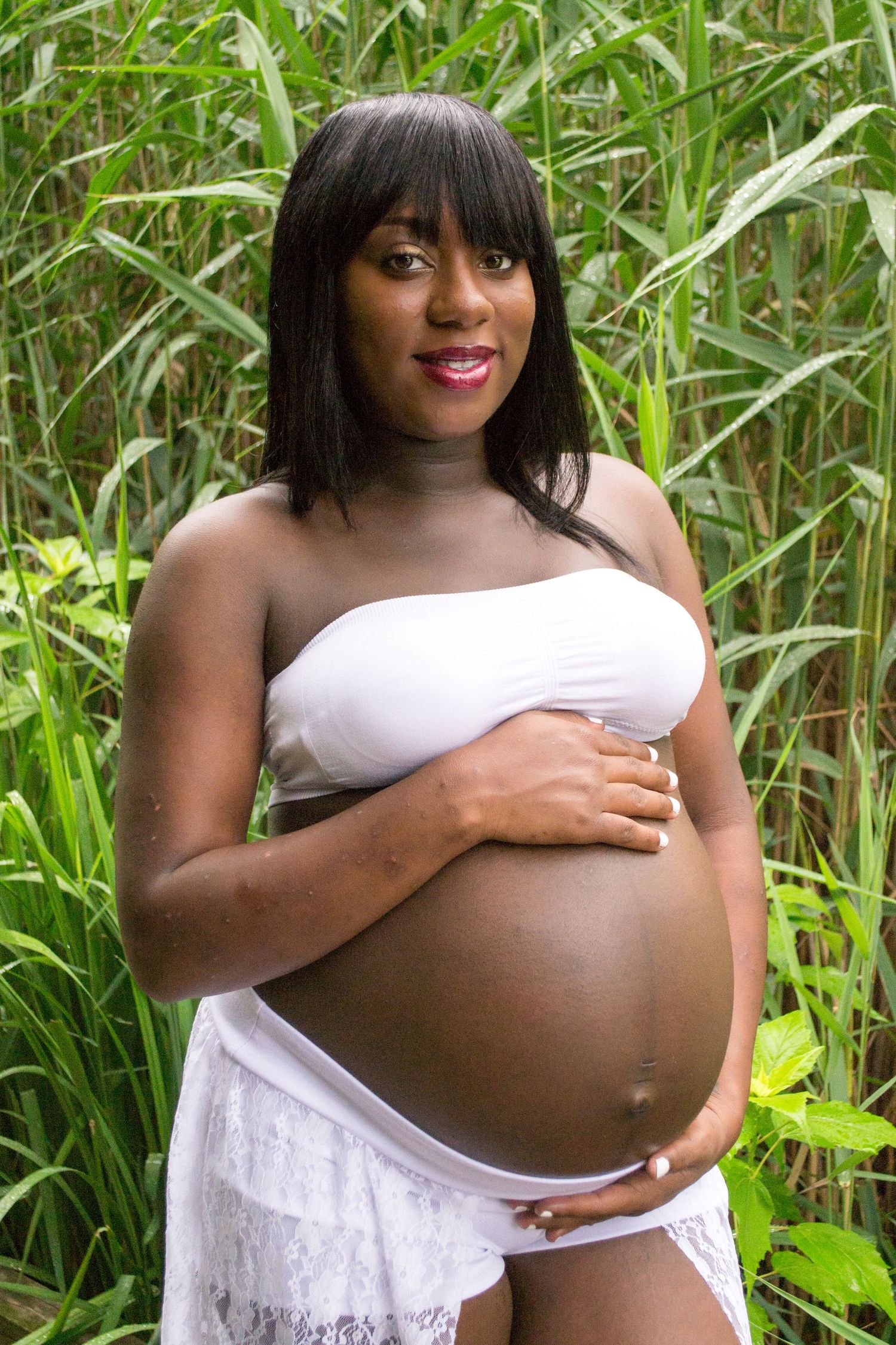 Pregnant Black woman in white bandeau top holding baby bump outdoors among tall green reeds.