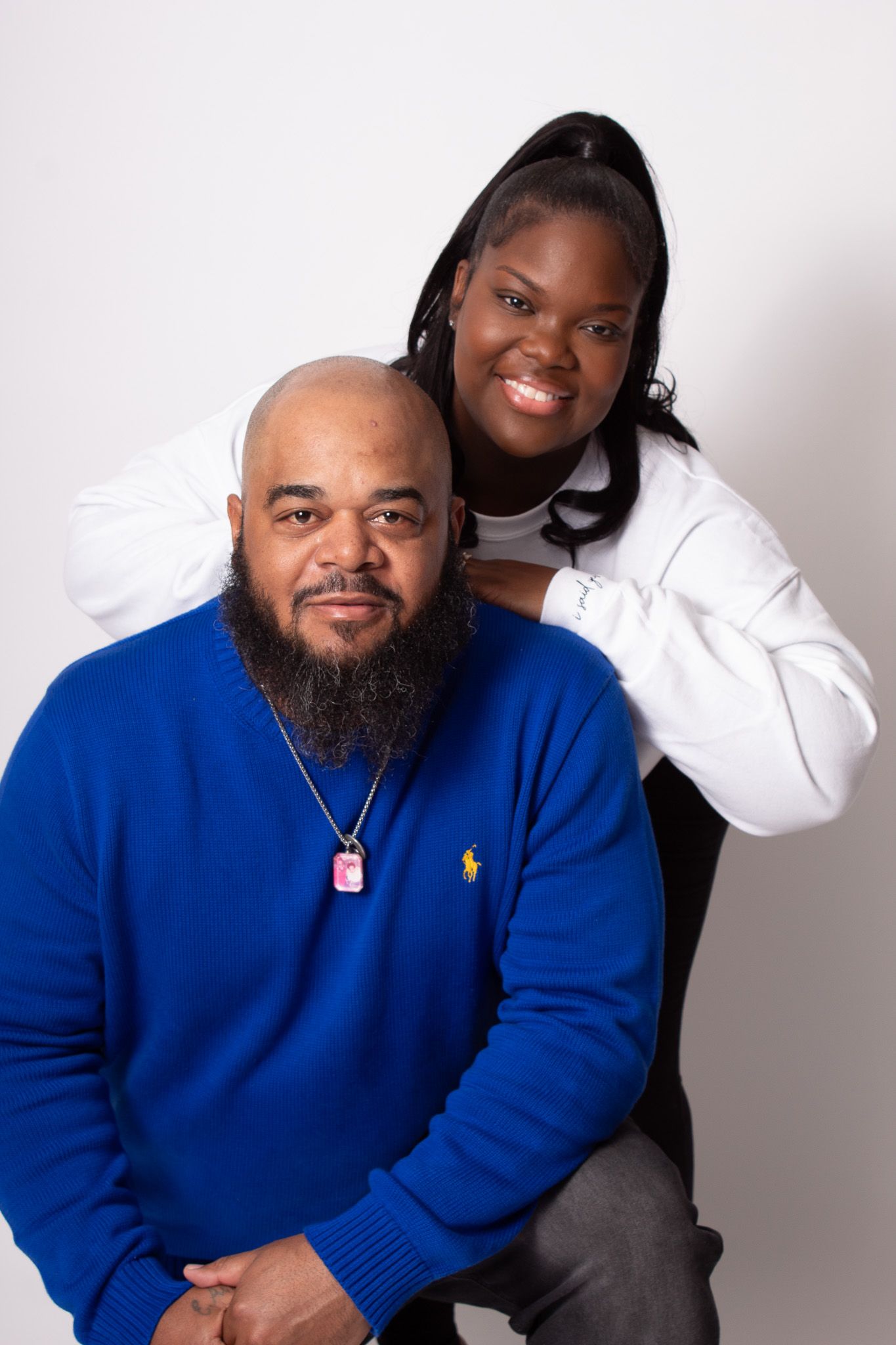 A bald bearded man in a blue Polo sweater poses with a smiling woman in a white hoodie against a white background.