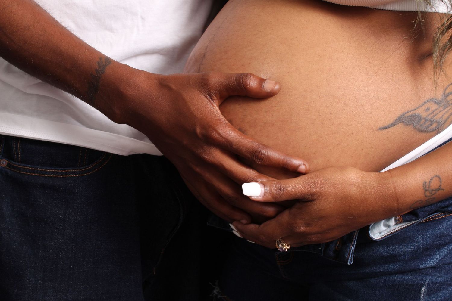 Close-up of a couple holding a pregnant woman's bare baby bump, showing a tattoo on her side.