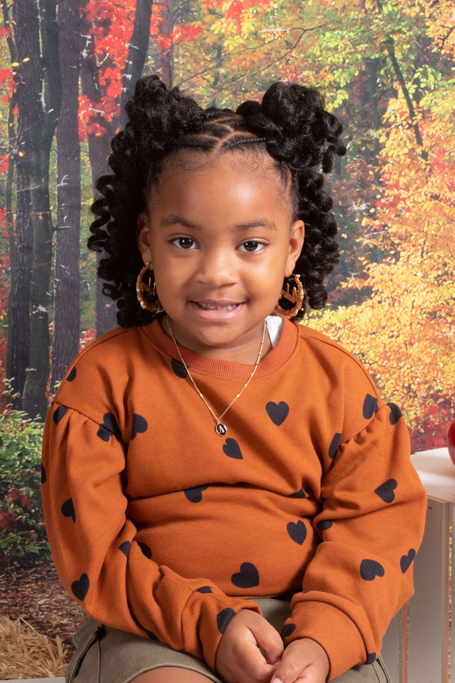 Young girl with curly puff hairstyle wearing orange heart-print sweatshirt smiling before autumn foliage backdrop.
