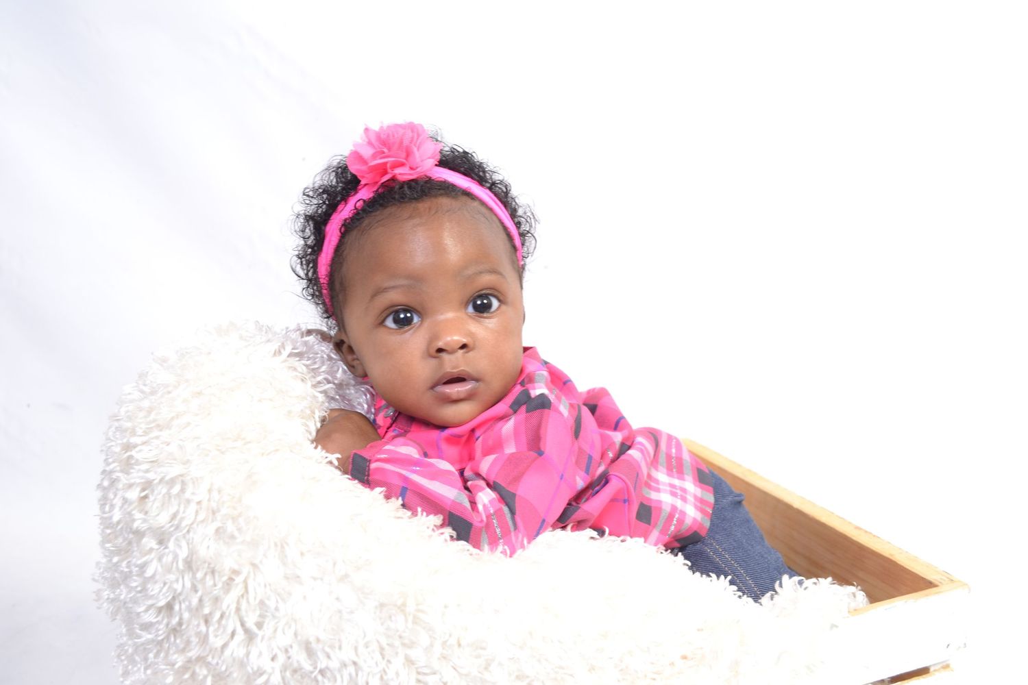 An adorable baby girl in pink outfit with floral headband sits in a wooden box lined with fluffy white fur.