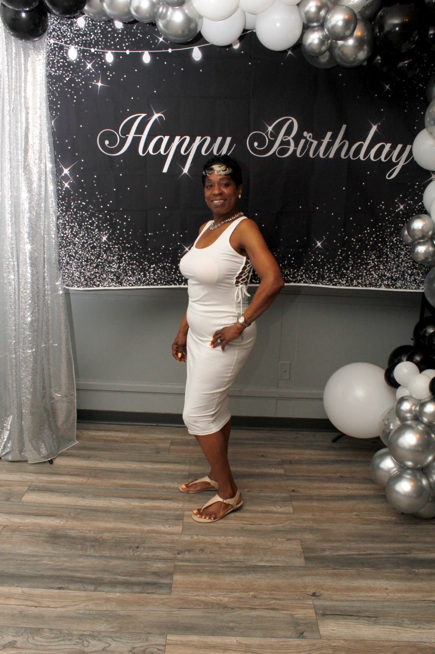 Woman in white dress posing at a black and silver Happy Birthday party with balloon arch decorations.