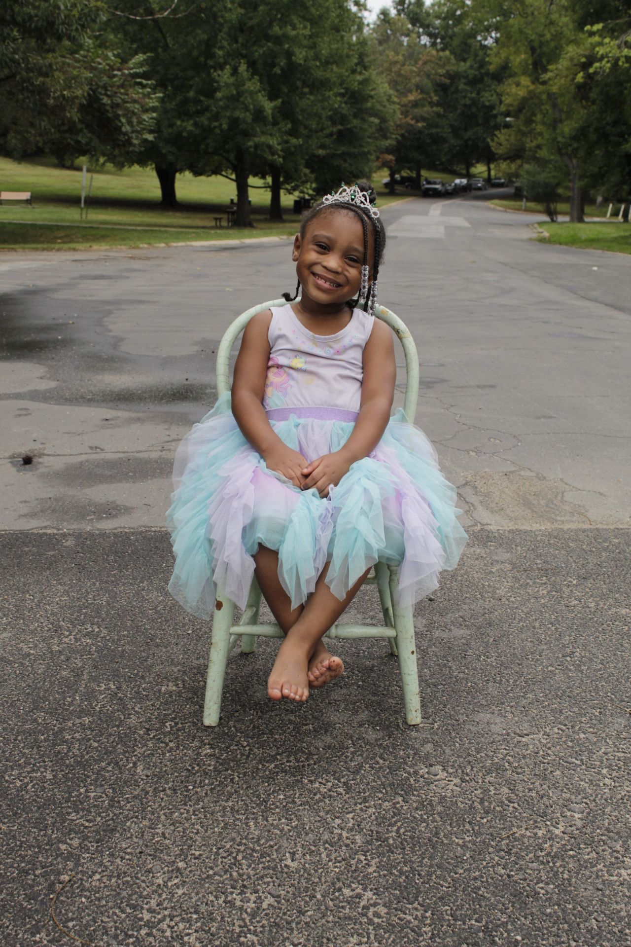 A young girl in a pastel tutu dress and tiara smiles sweetly while seated in a chair outdoors.