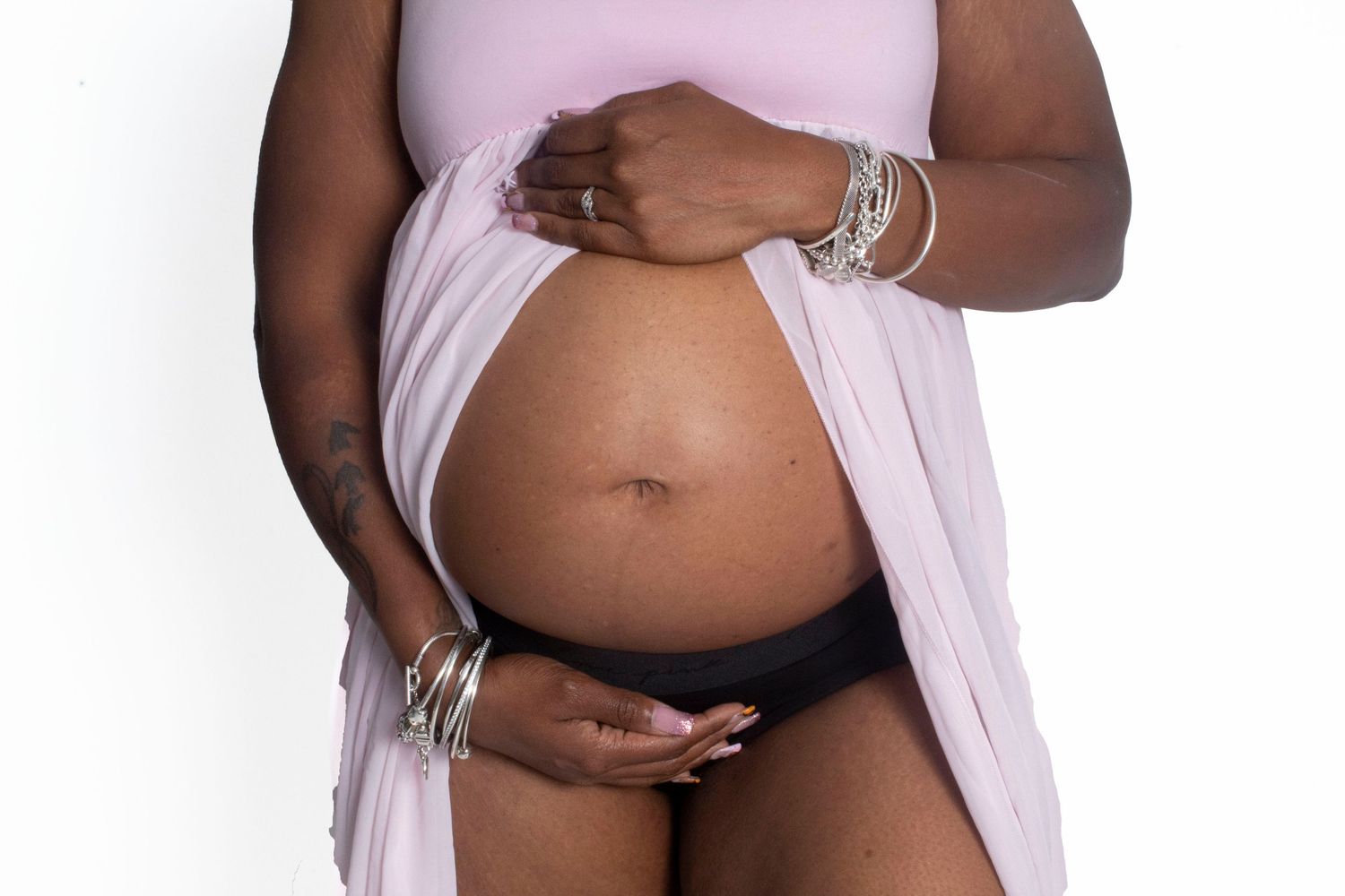 Close-up of pregnant Black woman in pink top and black underwear holding baby bump with silver bangles.
