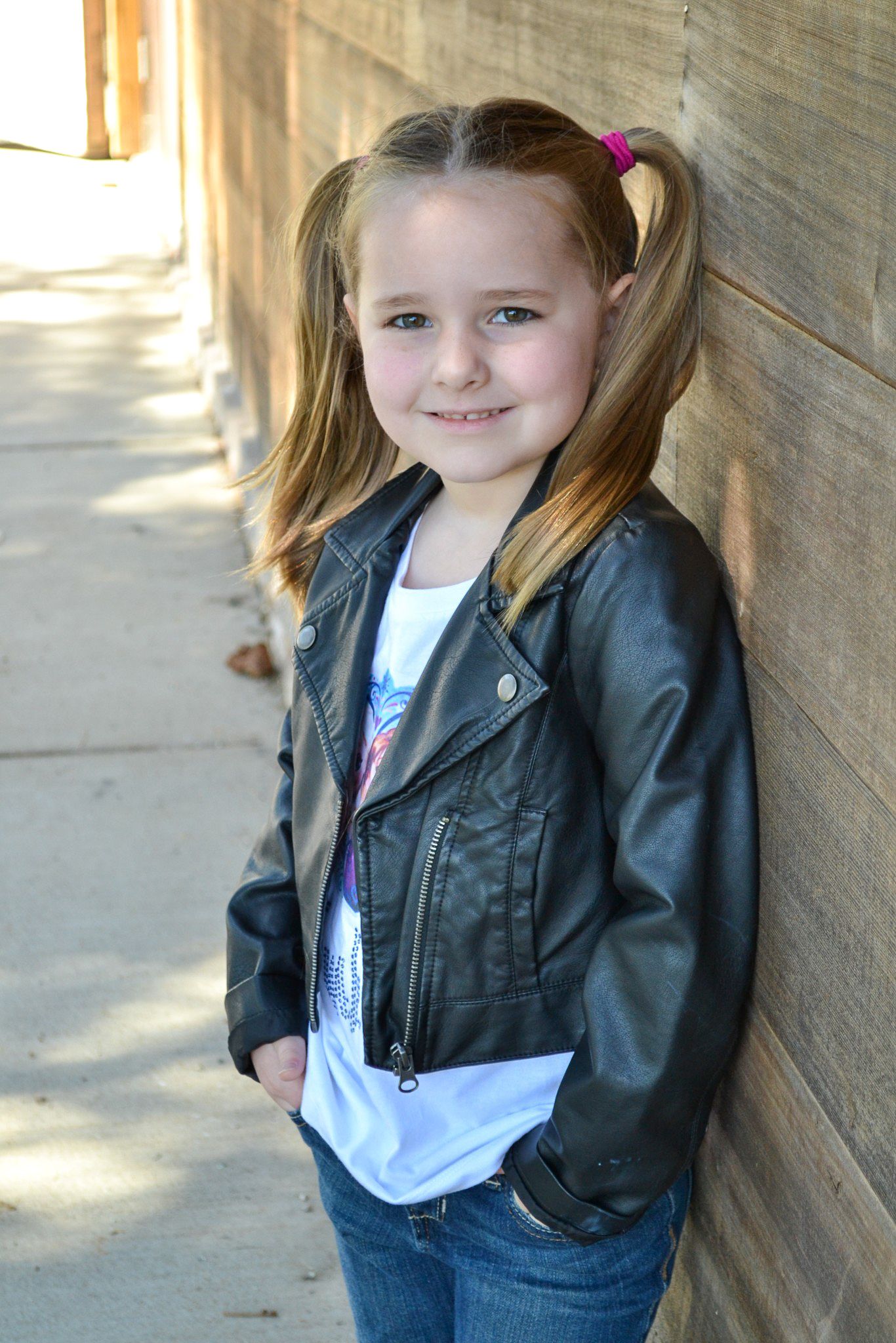 Young girl in black leather jacket and pigtails smiles while leaning against a wooden wall outdoors.