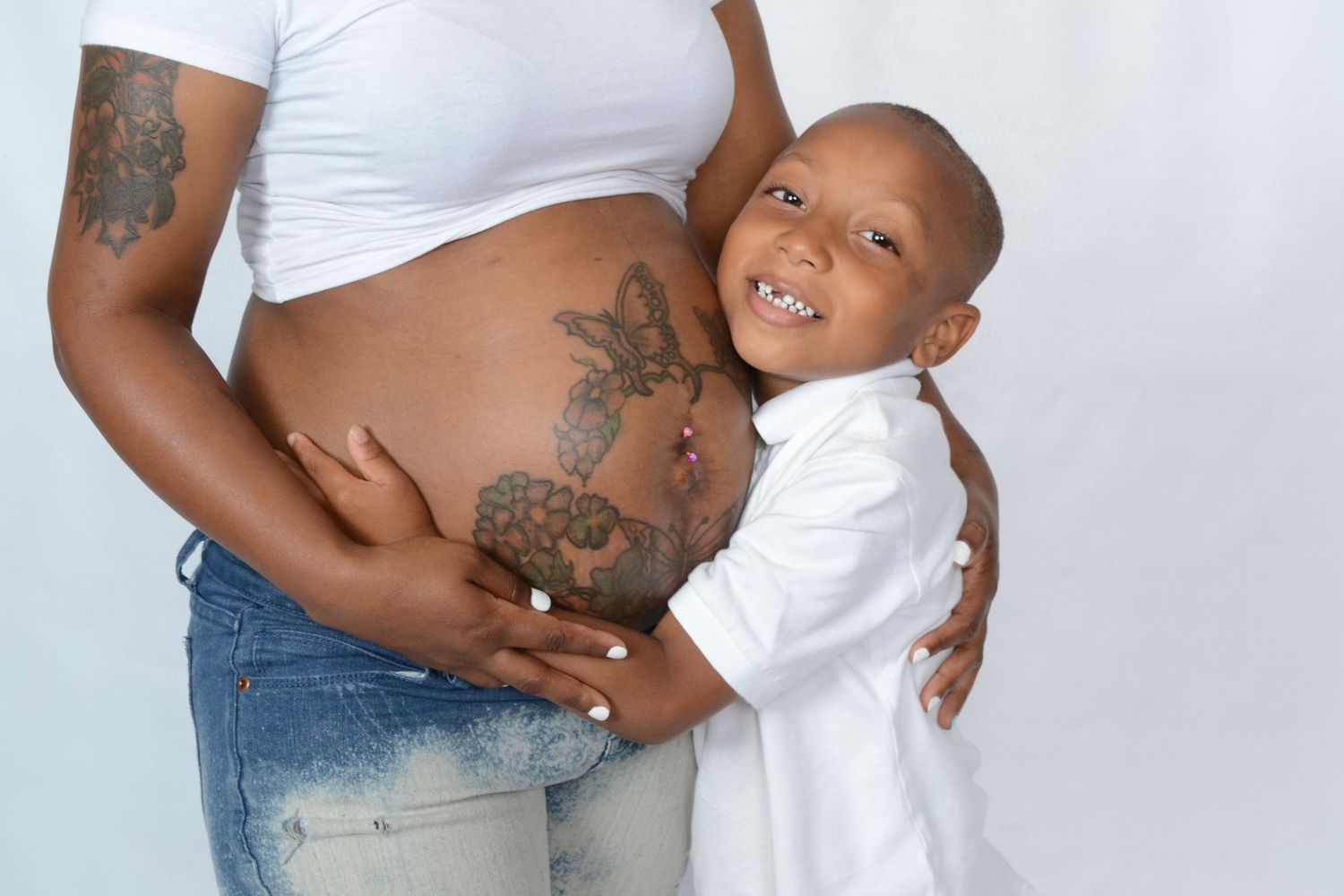 Young boy hugs pregnant mothers tattooed belly while smiling at camera against white background.