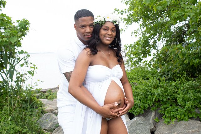 A couple in white poses outdoors for a maternity photo, the woman wearing a floral crown and cradling her baby bump.