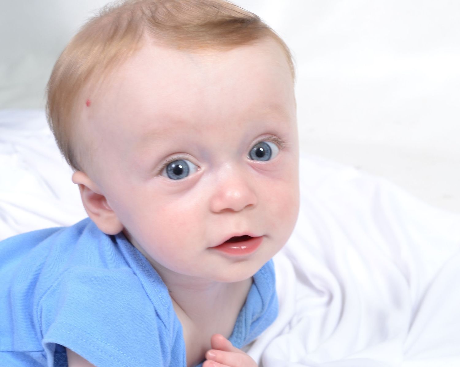 Close-up portrait of a blue-eyed baby boy in blue shirt lying on white blanket.