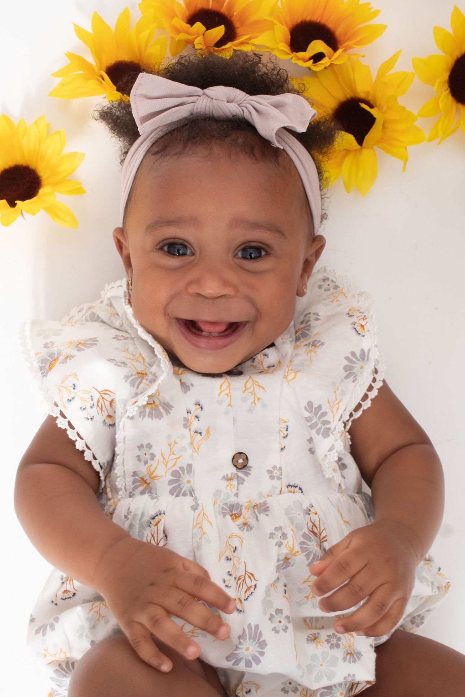 Joyful baby girl in floral dress with bow headband smiles surrounded by sunflowers.
