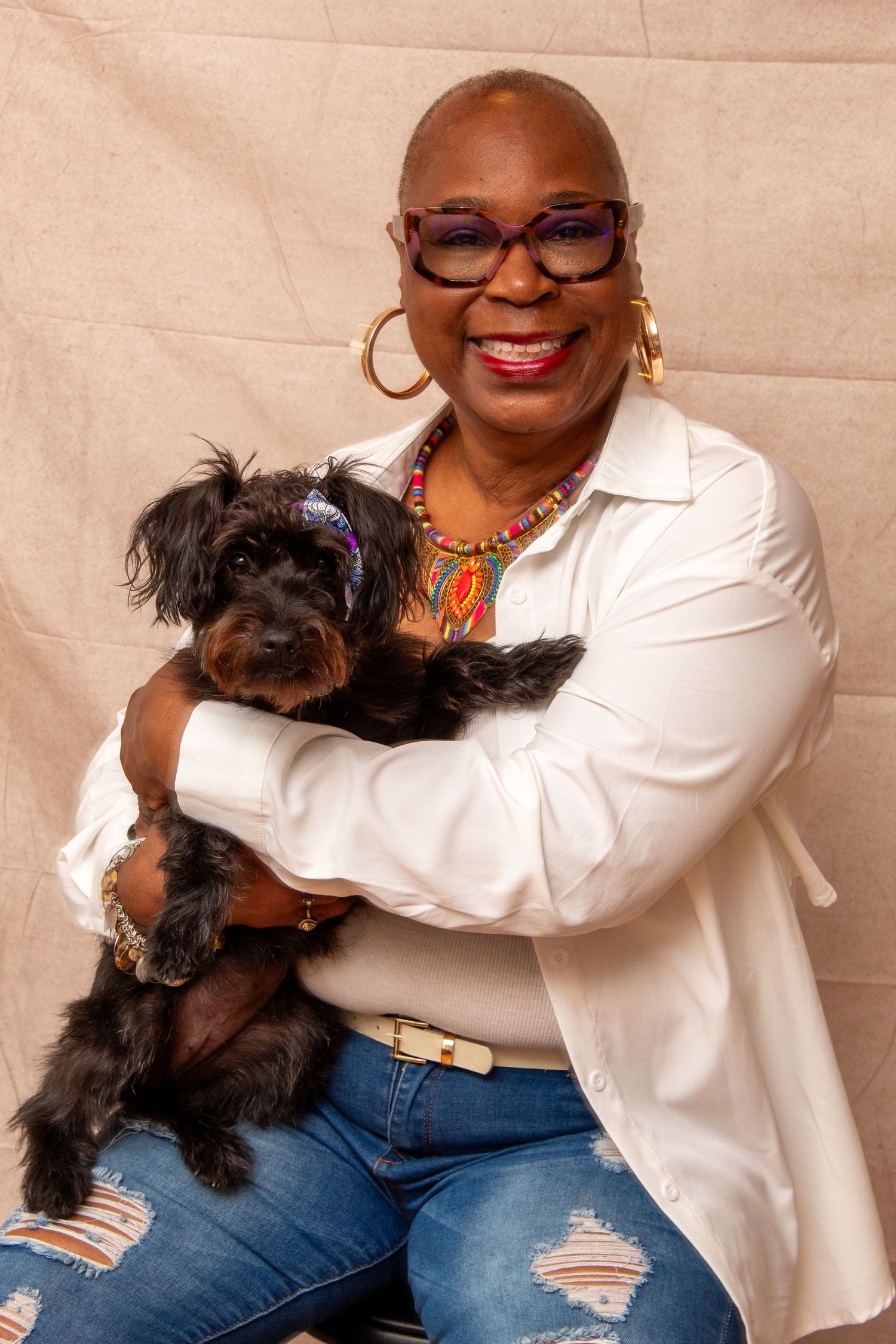 Smiling woman with glasses and colorful necklace holds a small black dog in a studio portrait.