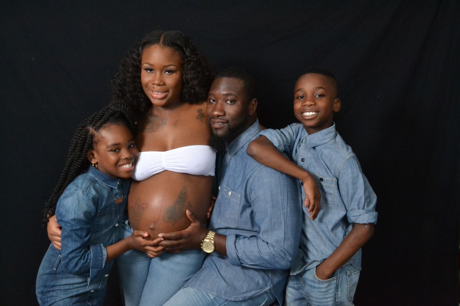 A joyful family maternity photo with a pregnant woman in white, her partner, and two children in denim outfits on black background.