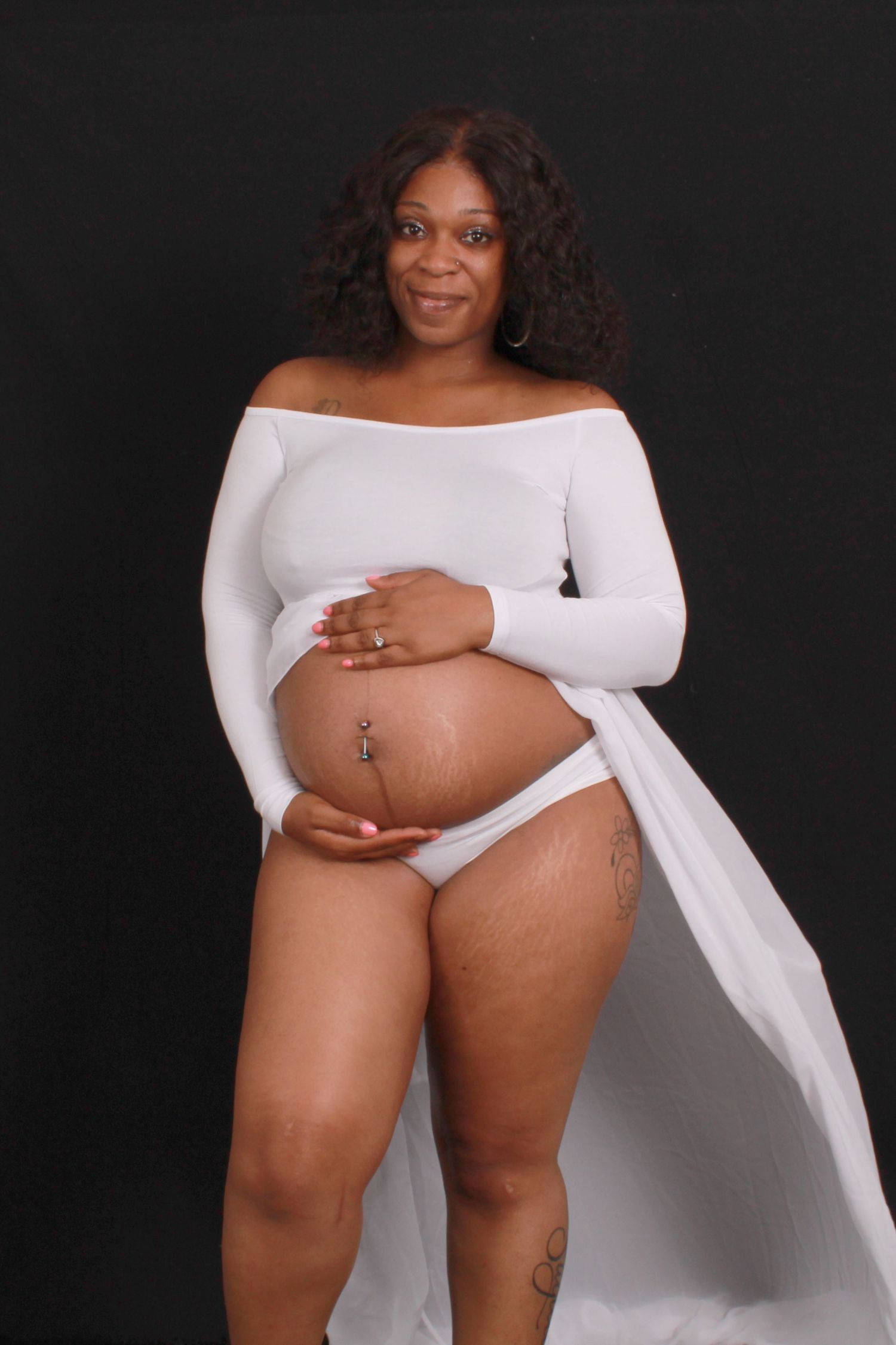 Smiling pregnant Black woman in white bodysuit and underwear showing baby bump with tattoos visible on legs.
