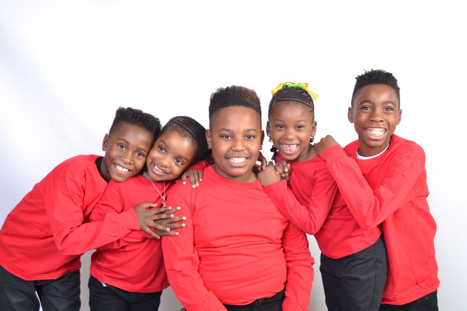 Five children in matching red shirts smile and hug together in a cheerful group portrait.