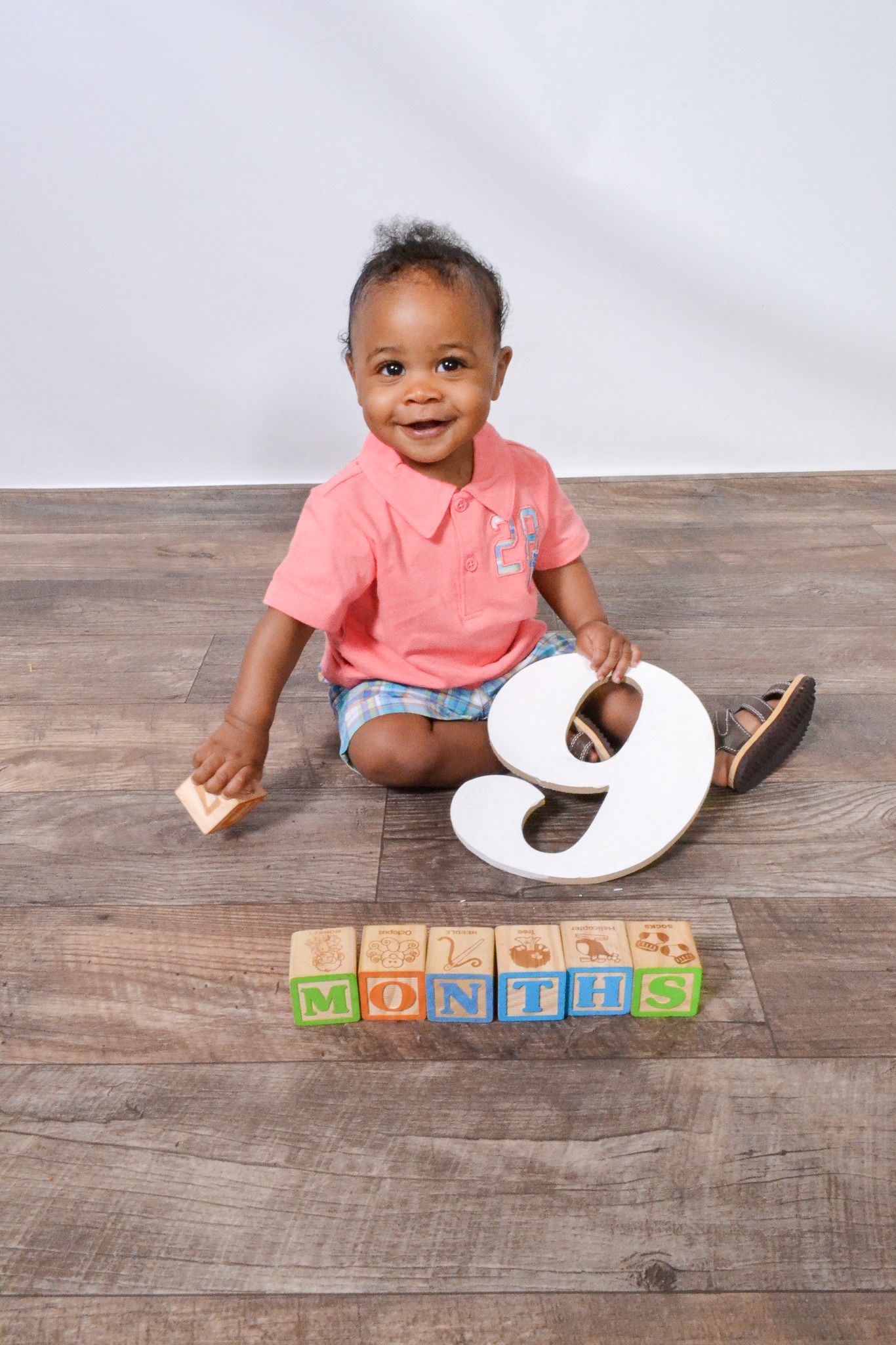 Smiling baby boy sitting on wood floor with alphabet blocks spelling 9 MONTHS for milestone photo.