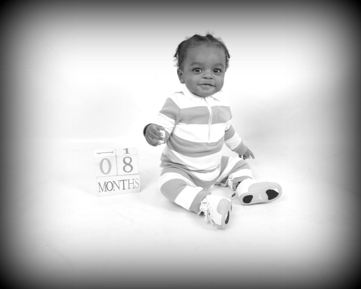 Black and white photo of a baby at 18 months old sitting up, wearing a striped outfit and panda shoes.