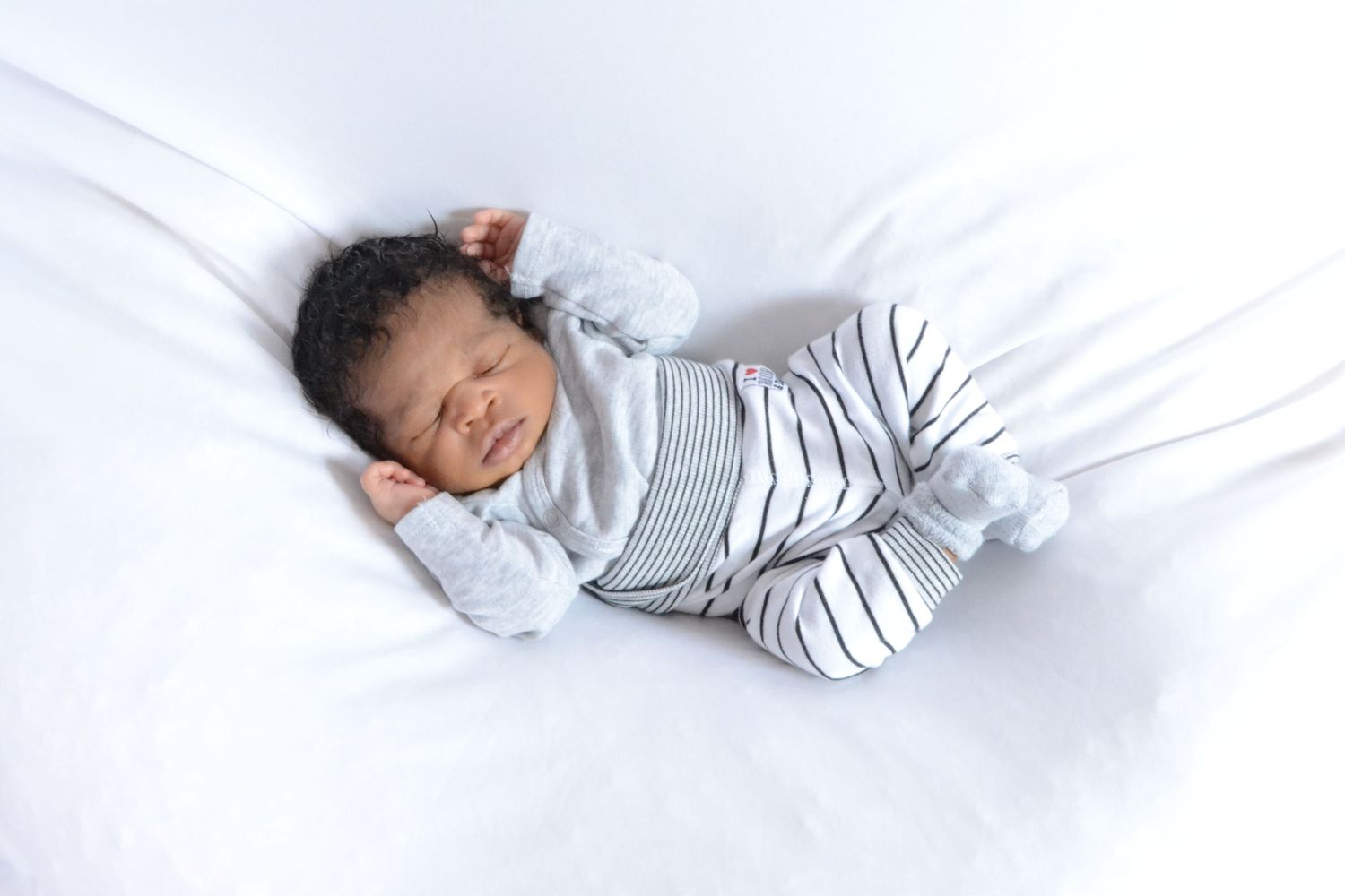 Sleeping newborn baby in striped gray outfit lying on white bedding.