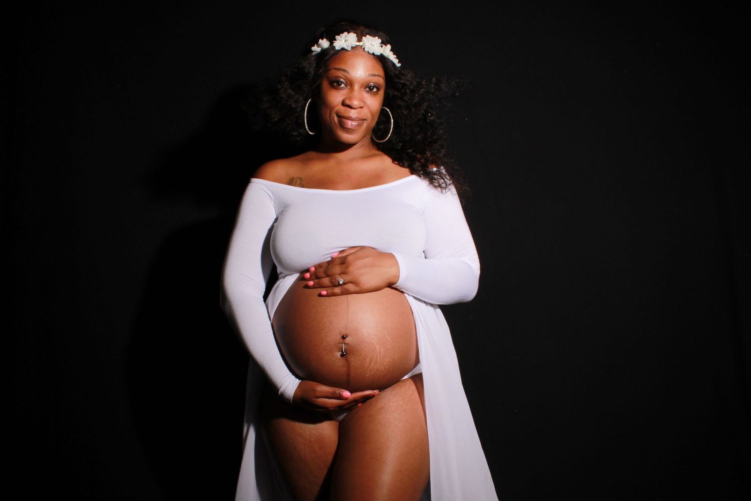 Pregnant Black woman wearing white floral crown and off-shoulder white top proudly displaying bare baby bump.