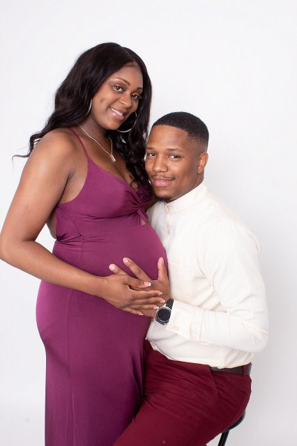 Pregnant woman in purple dress poses with partner kneeling beside her holding her baby bump in a studio.