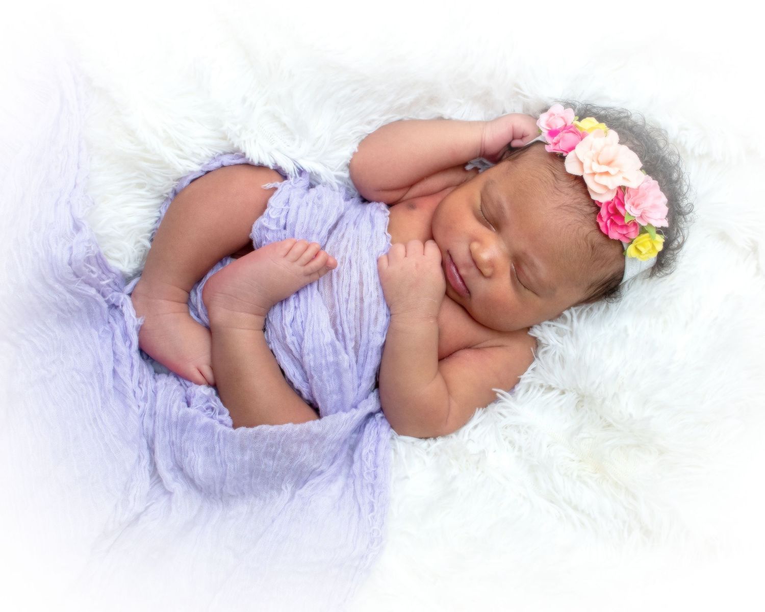 A peaceful newborn baby sleeps wrapped in lavender cloth with a colorful floral headband on a white fluffy blanket.