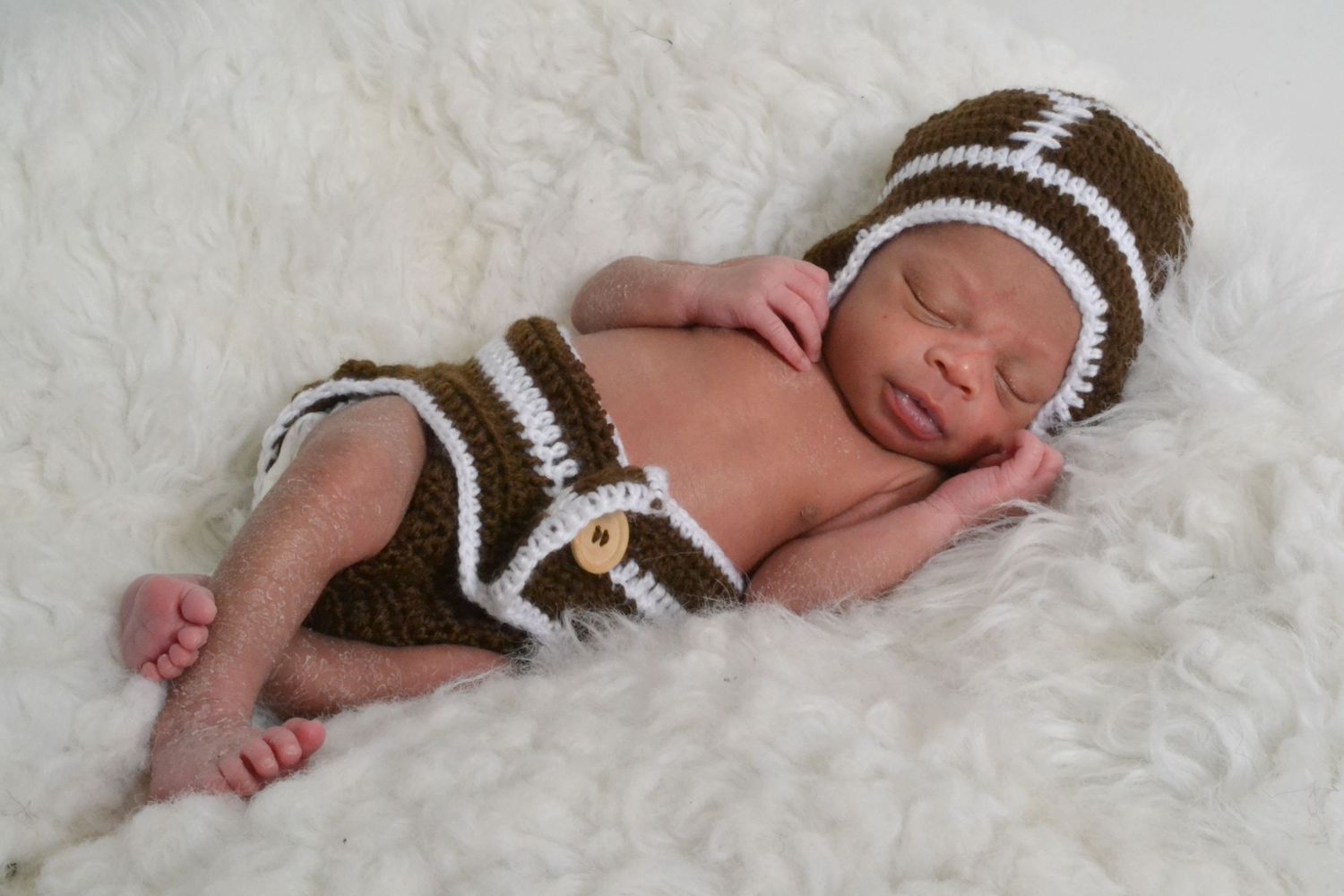 Newborn baby sleeping in crocheted football outfit and hat on white fluffy blanket.