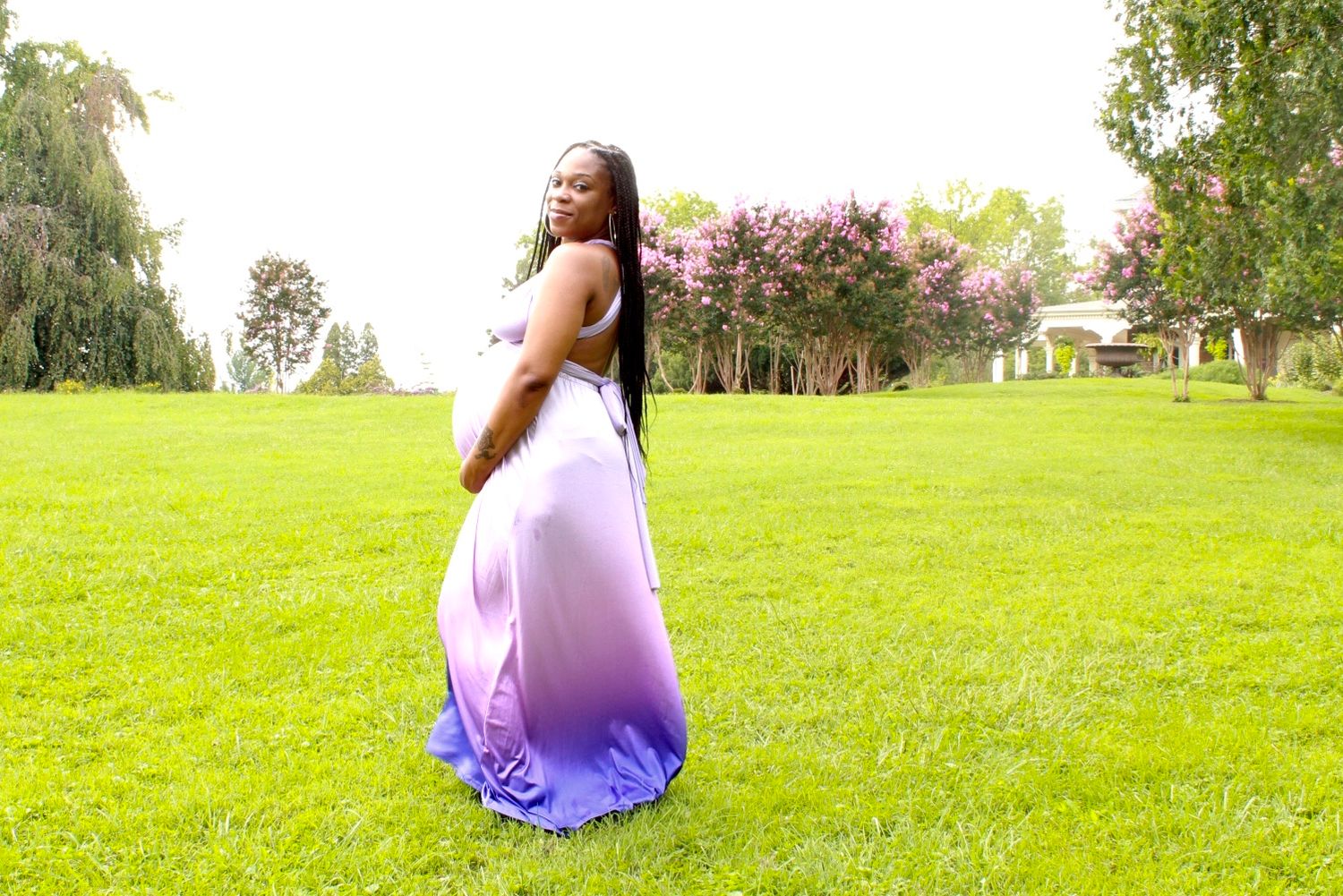 A pregnant woman in a flowing purple ombre gown poses gracefully on a lush green lawn outdoors.