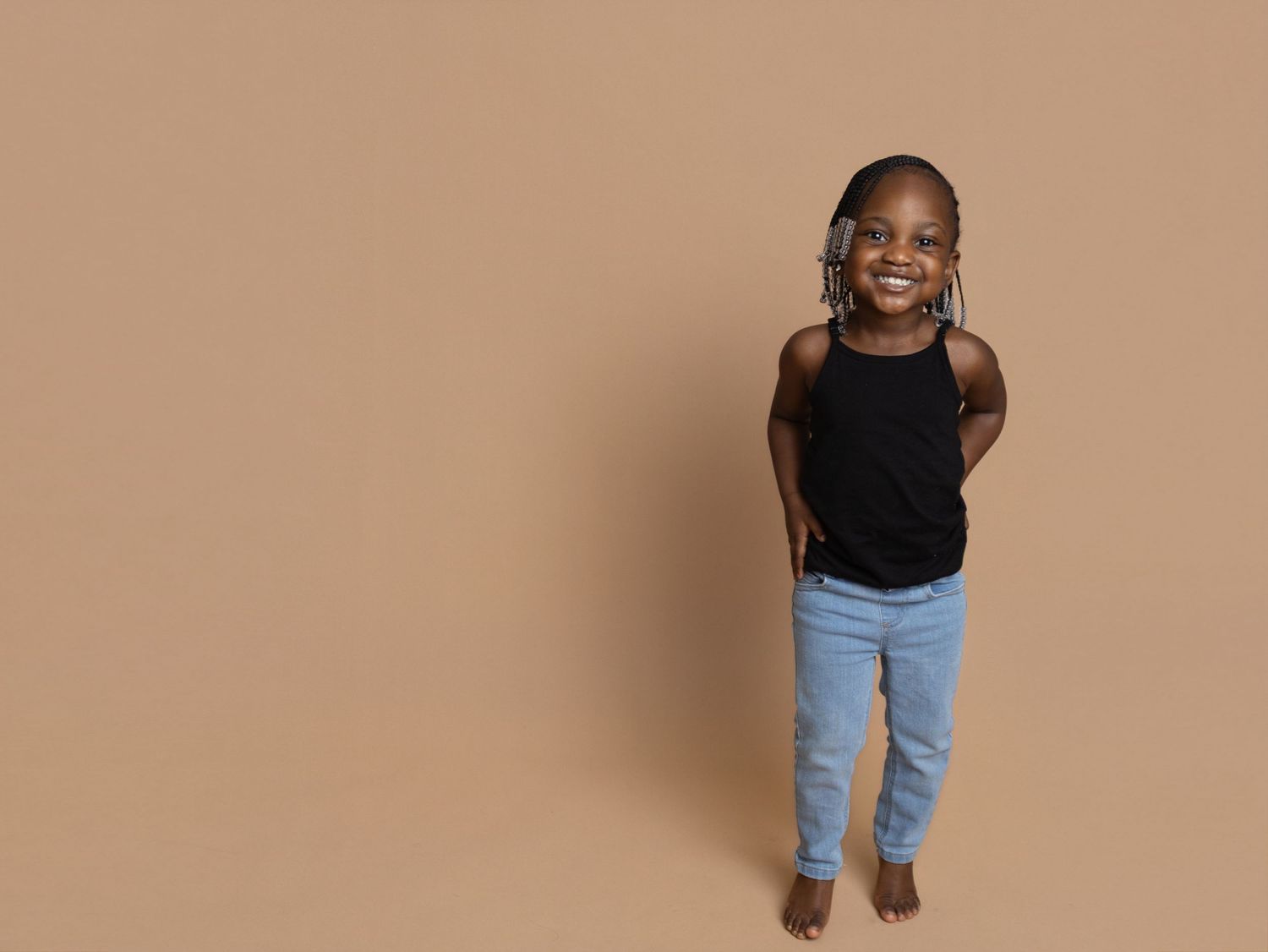 Smiling young Black girl in black tank top and blue jeans standing against a warm beige background.