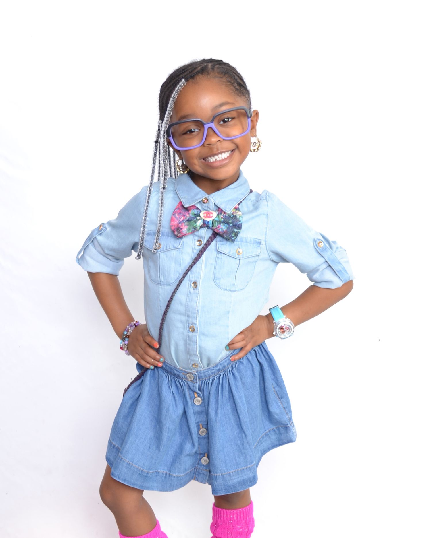 Stylish young girl in denim outfit with bow tie and purple glasses poses confidently smiling.