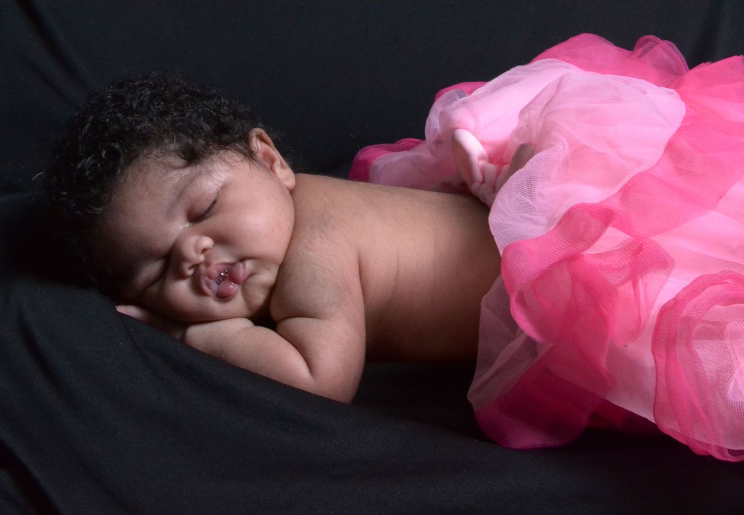 Sleeping newborn baby posed in a fluffy pink tutu on a dark background.