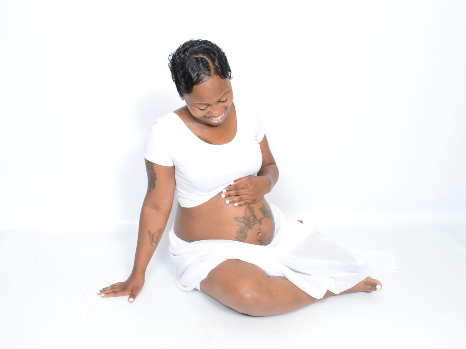 Pregnant woman in white outfit sits cross-legged on floor, lovingly cradling her baby bump.
