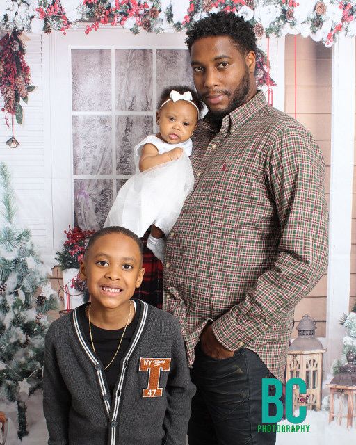 A father holds his baby girl while his young son smiles in a Christmas-themed family portrait.