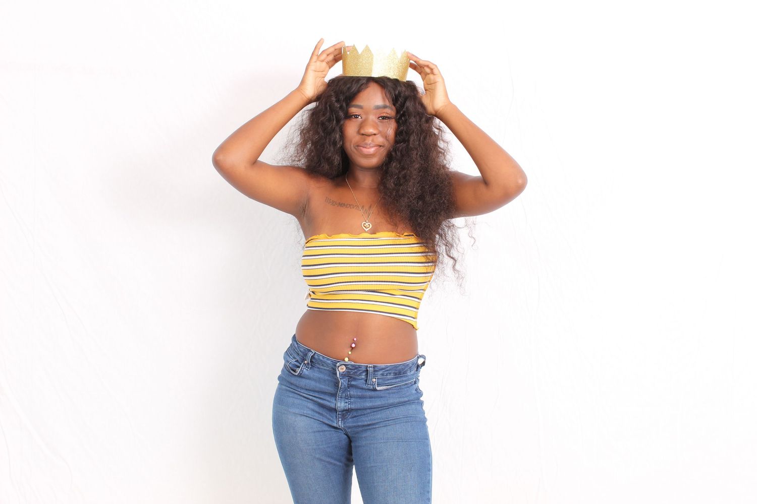 Young Black woman wearing yellow striped crop top and jeans, placing gold crown on her curly hair against white background.
