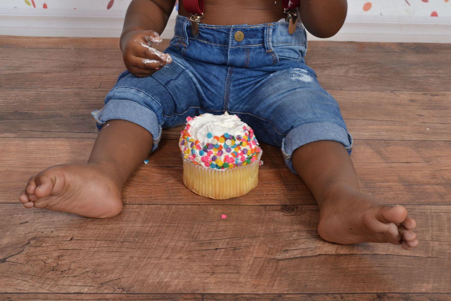 Toddler sitting on wood floor with colorful sprinkle birthday smash cake during first birthday celebration.
