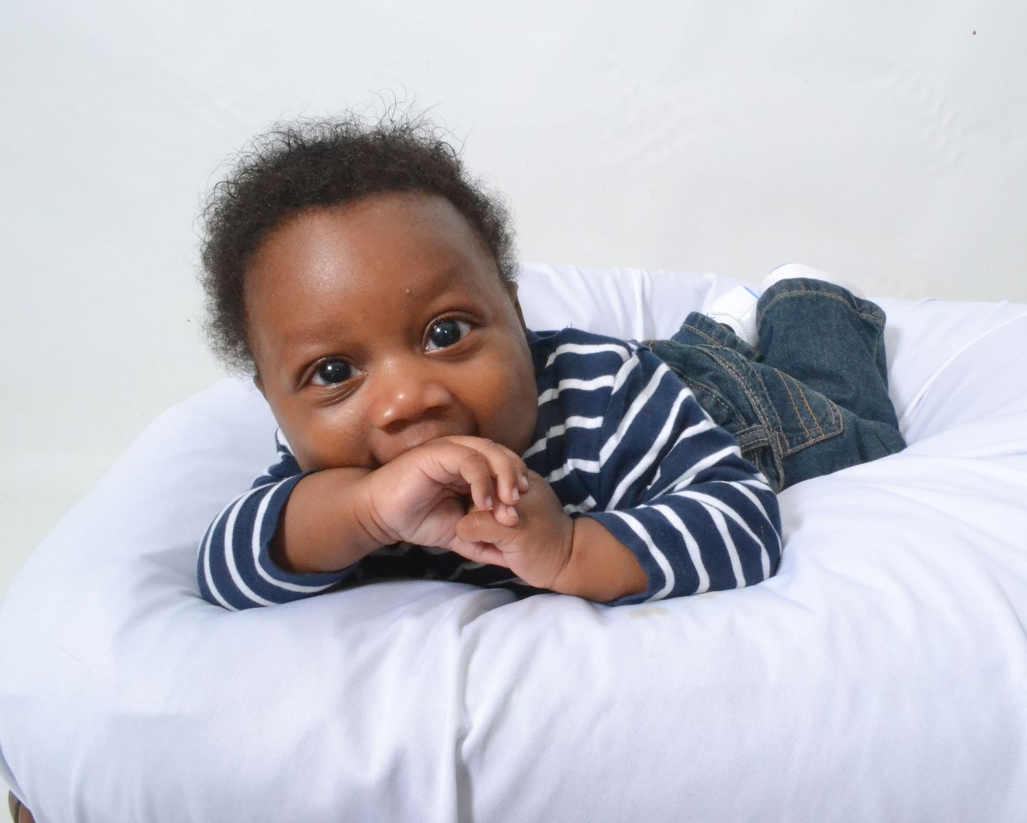 Adorable Black baby boy lying on white cushion in striped shirt and jeans, smiling with hands clasped.
