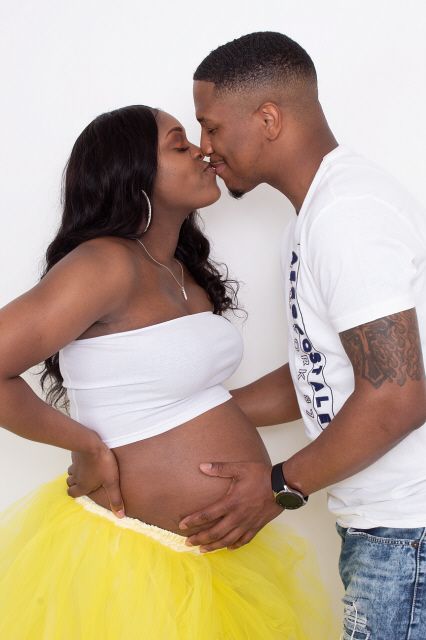 Pregnant couple sharing a kiss during maternity photoshoot, woman in white tube top and yellow tutu skirt.