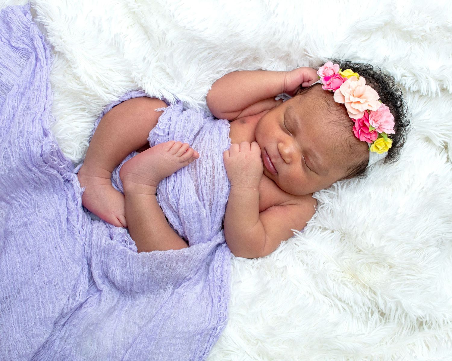 Sleeping newborn baby wrapped in purple cloth with floral headband on white fluffy blanket.