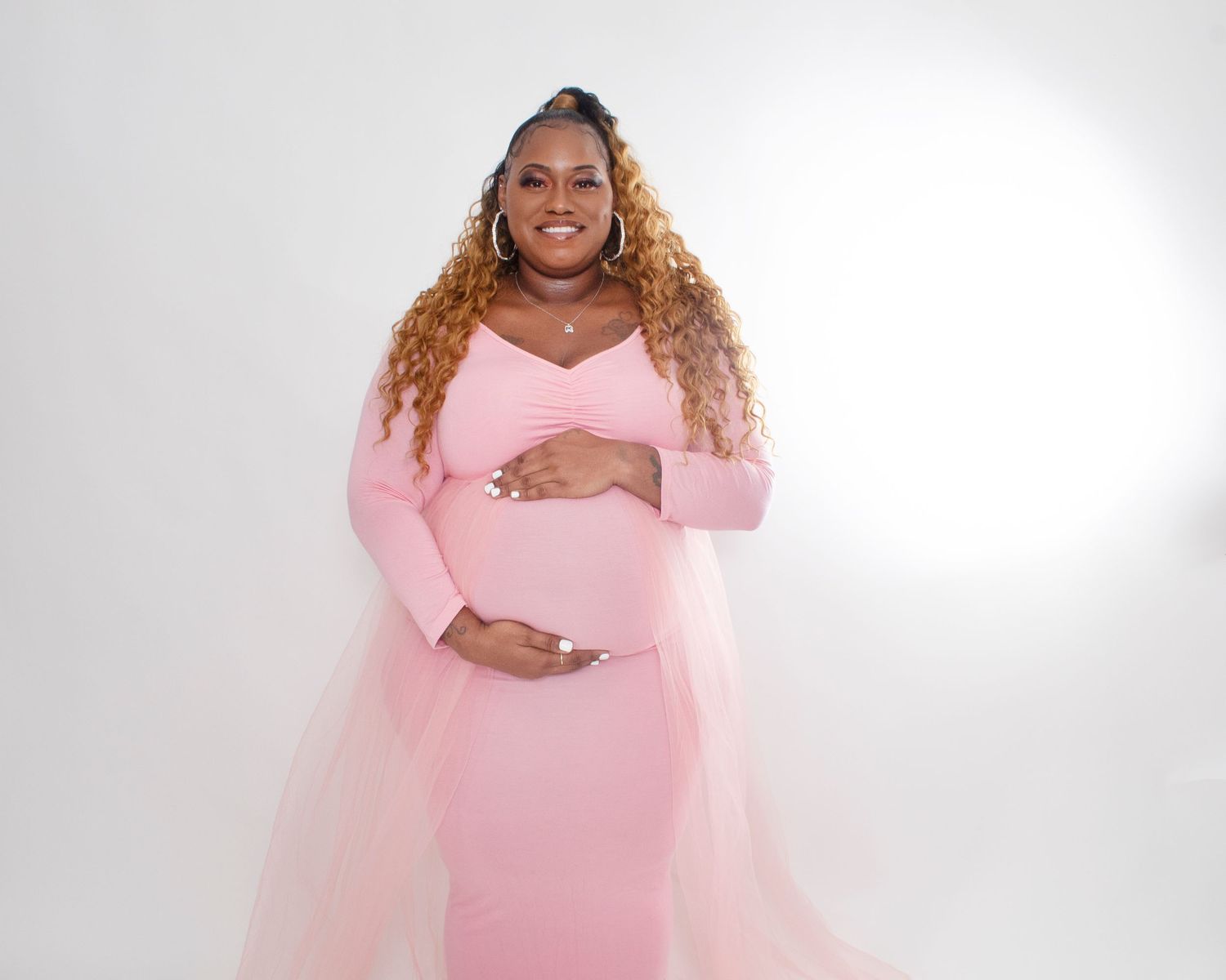 Pregnant woman in pink maternity gown with tulle train posing gracefully against white background.