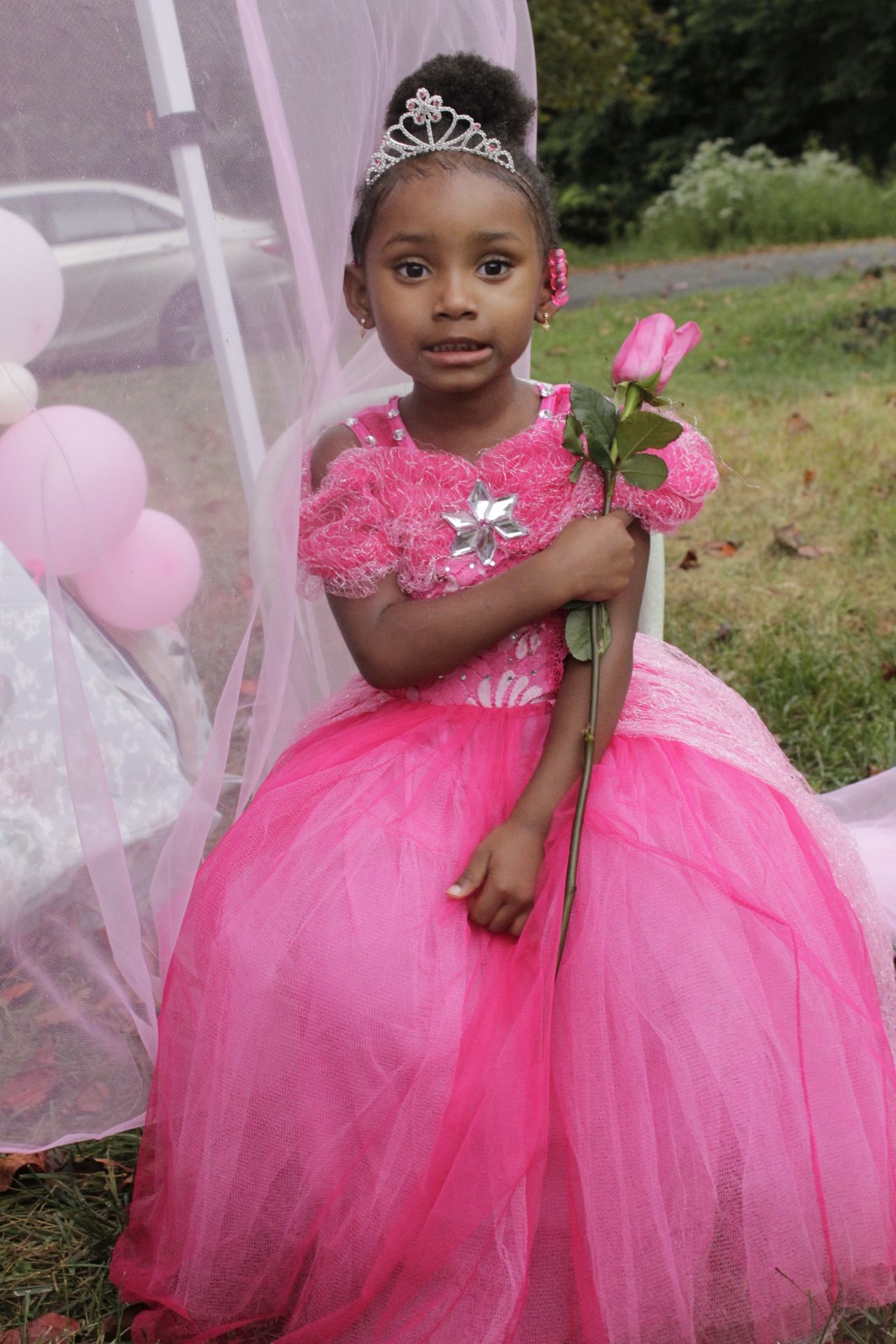 A young girl poses elegantly in a pink princess gown and tiara holding a pink rose.