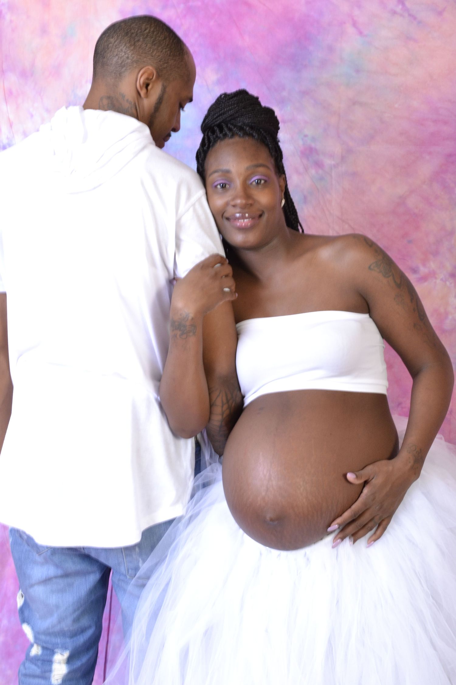 Pregnant woman in white strapless top and tulle skirt poses with partner against colorful pink backdrop.