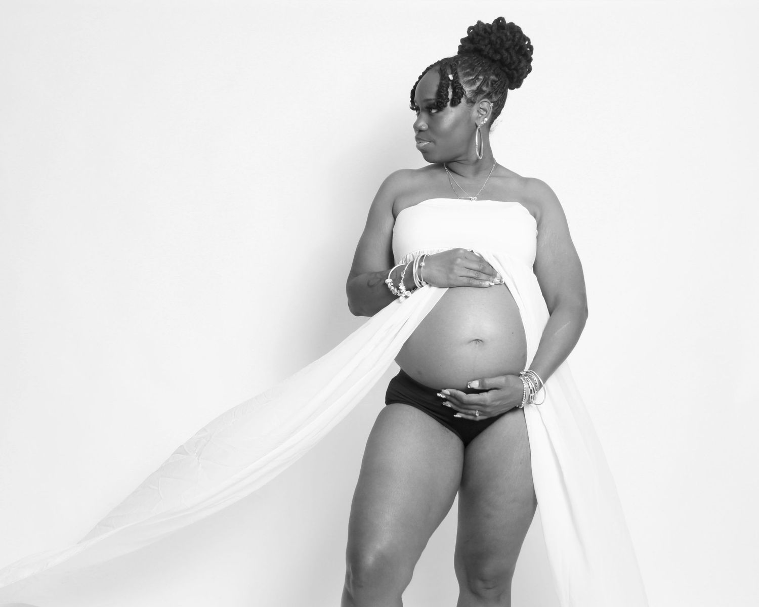Black and white maternity photo of a pregnant woman in white wrap and sunglasses posing gracefully in a studio.