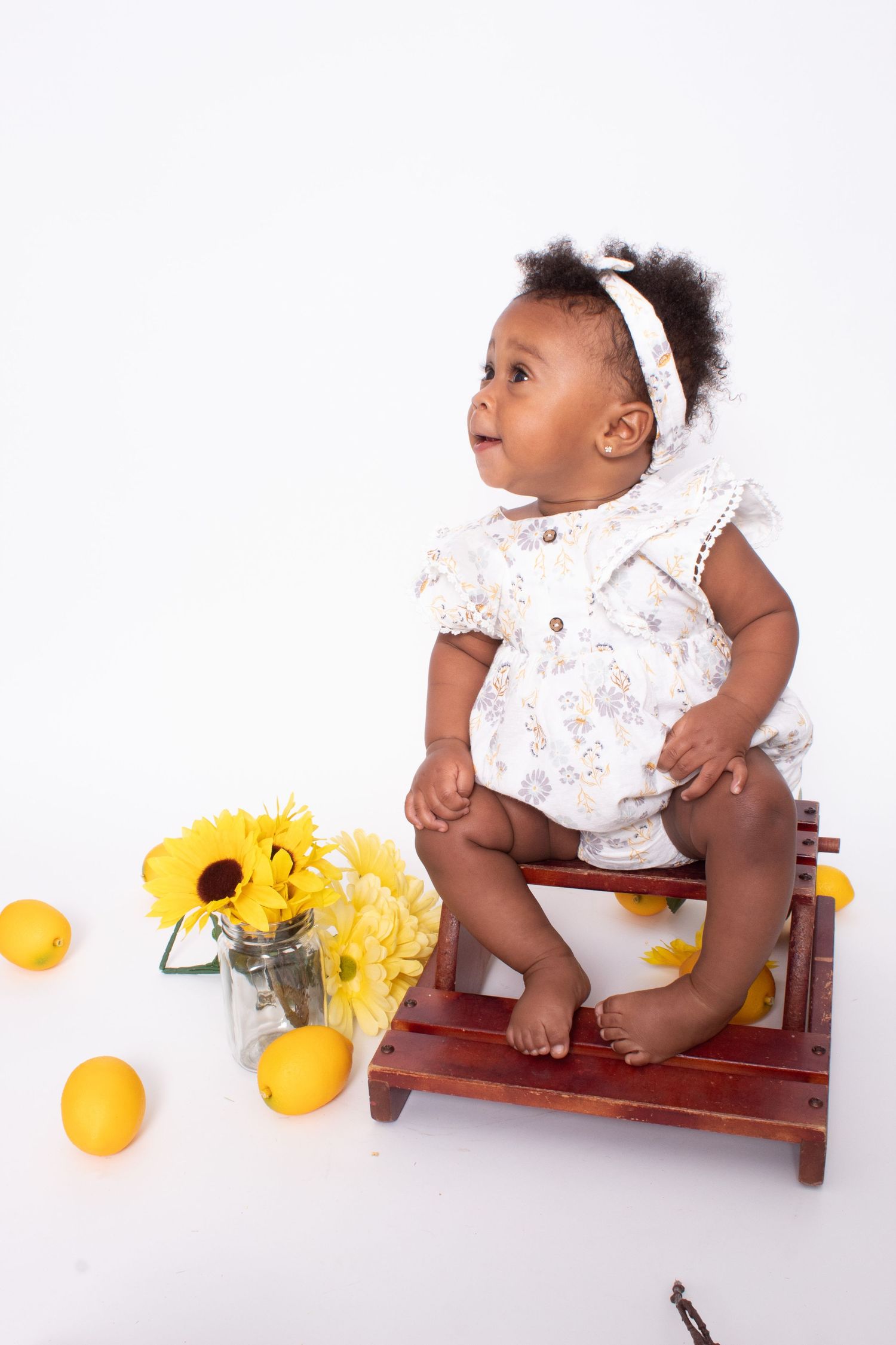 Adorable baby girl in floral romper sitting on wooden stool with sunflowers and lemons on white background.