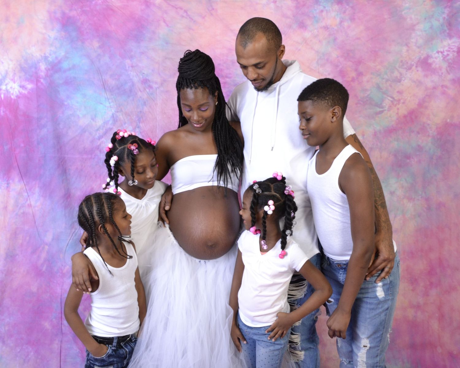 A pregnant woman in white poses with her partner and children against a colorful studio backdrop for a maternity photo.