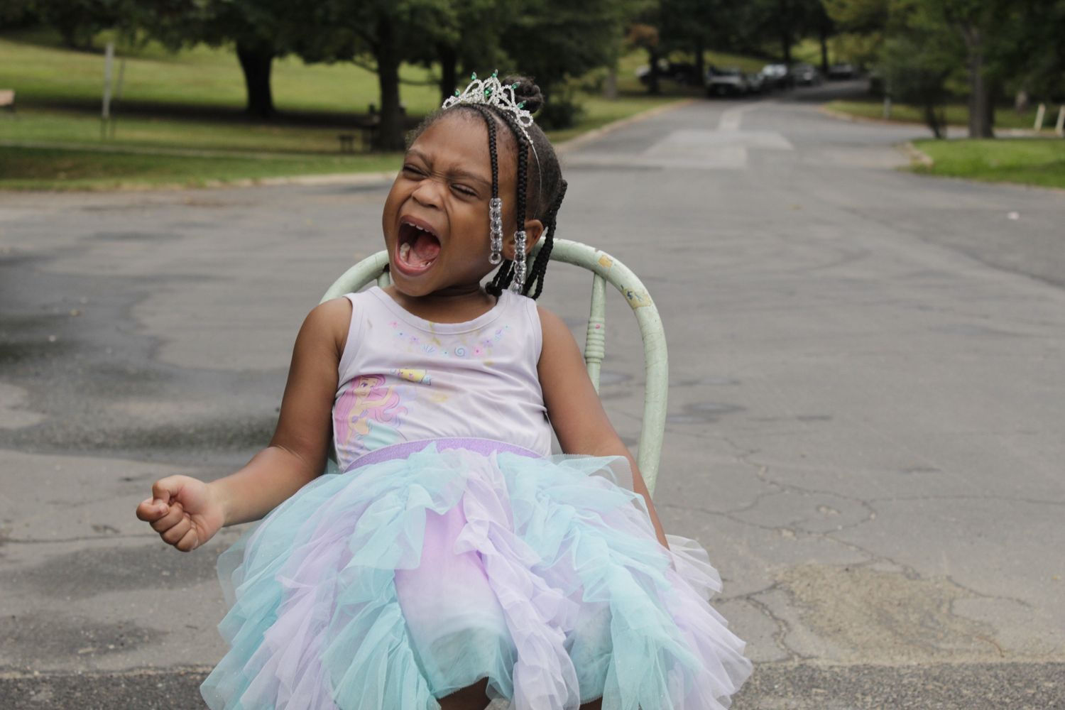 A young girl in a pastel tutu dress and tiara laughs joyfully while seated outdoors.