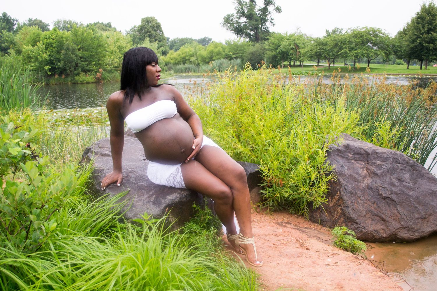 A pregnant woman in white sits on rocks by a serene pond surrounded by lush green foliage in an outdoor maternity shoot.