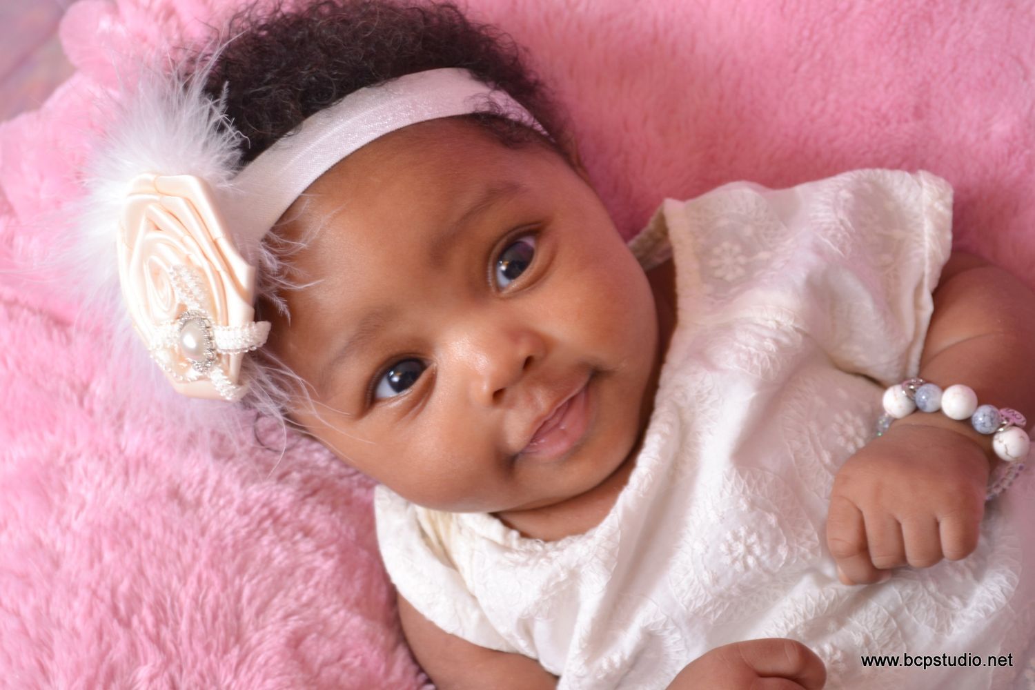 Adorable smiling newborn baby girl wearing a white dress and floral headband lying on a pink fluffy blanket.