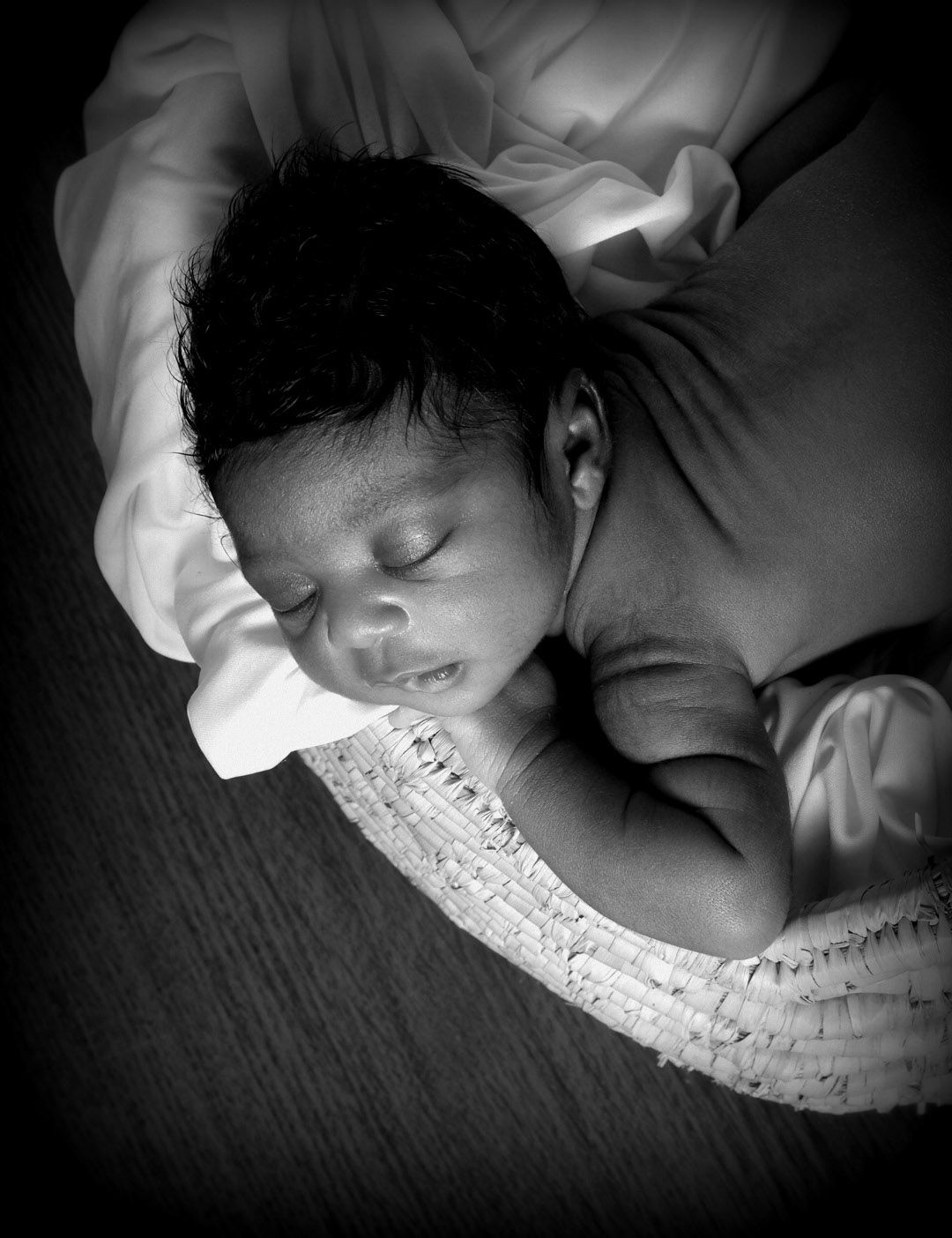 Black and white photo of a sleeping newborn baby with dark hair resting peacefully in a woven basket.