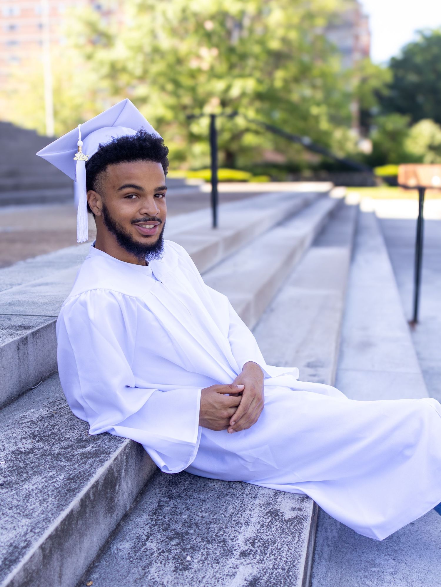 Young man in white graduation cap and gown sitting on stone steps, smiling outdoors on a sunny day.