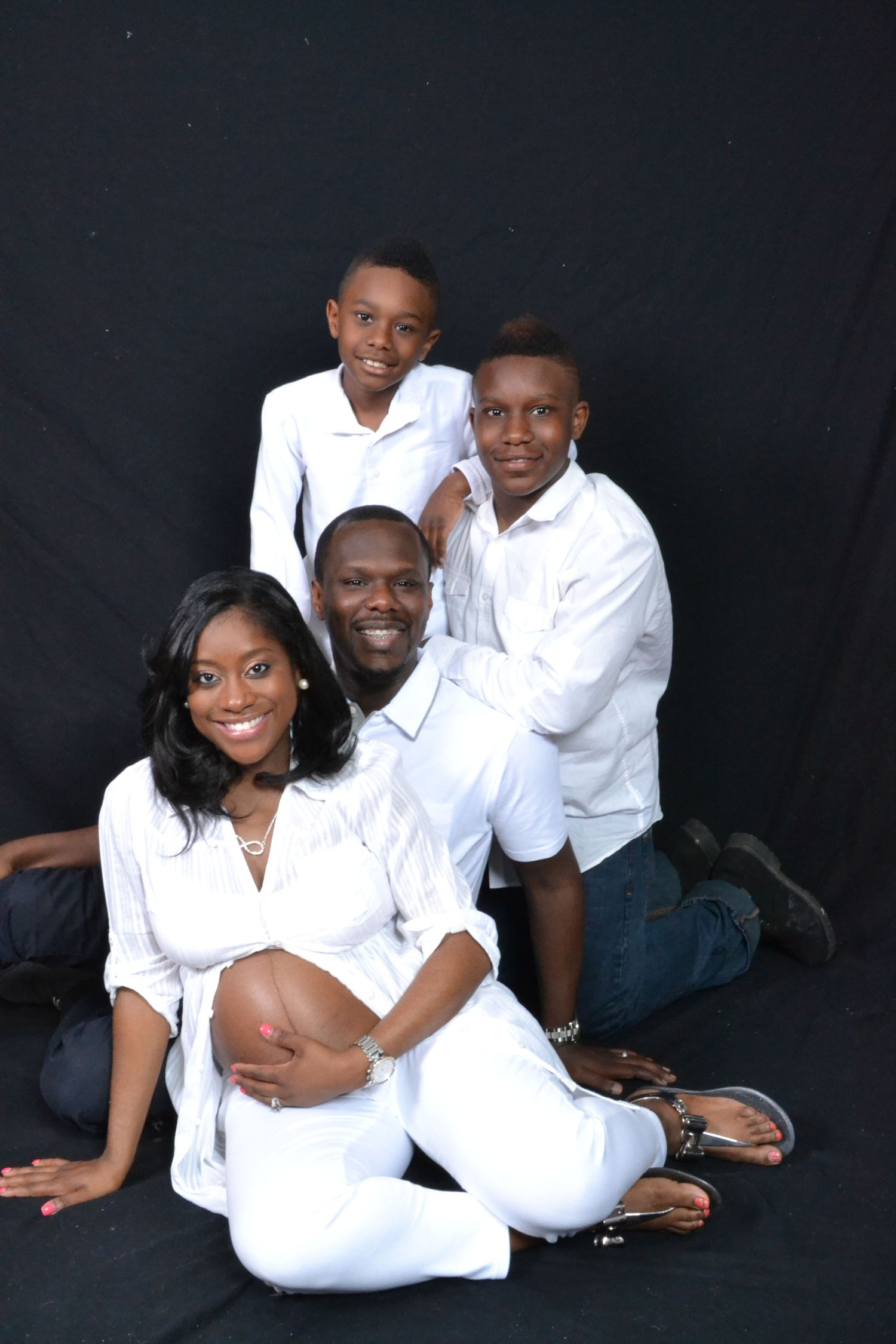 Family of four in matching white outfits poses for a maternity portrait against a black studio backdrop.