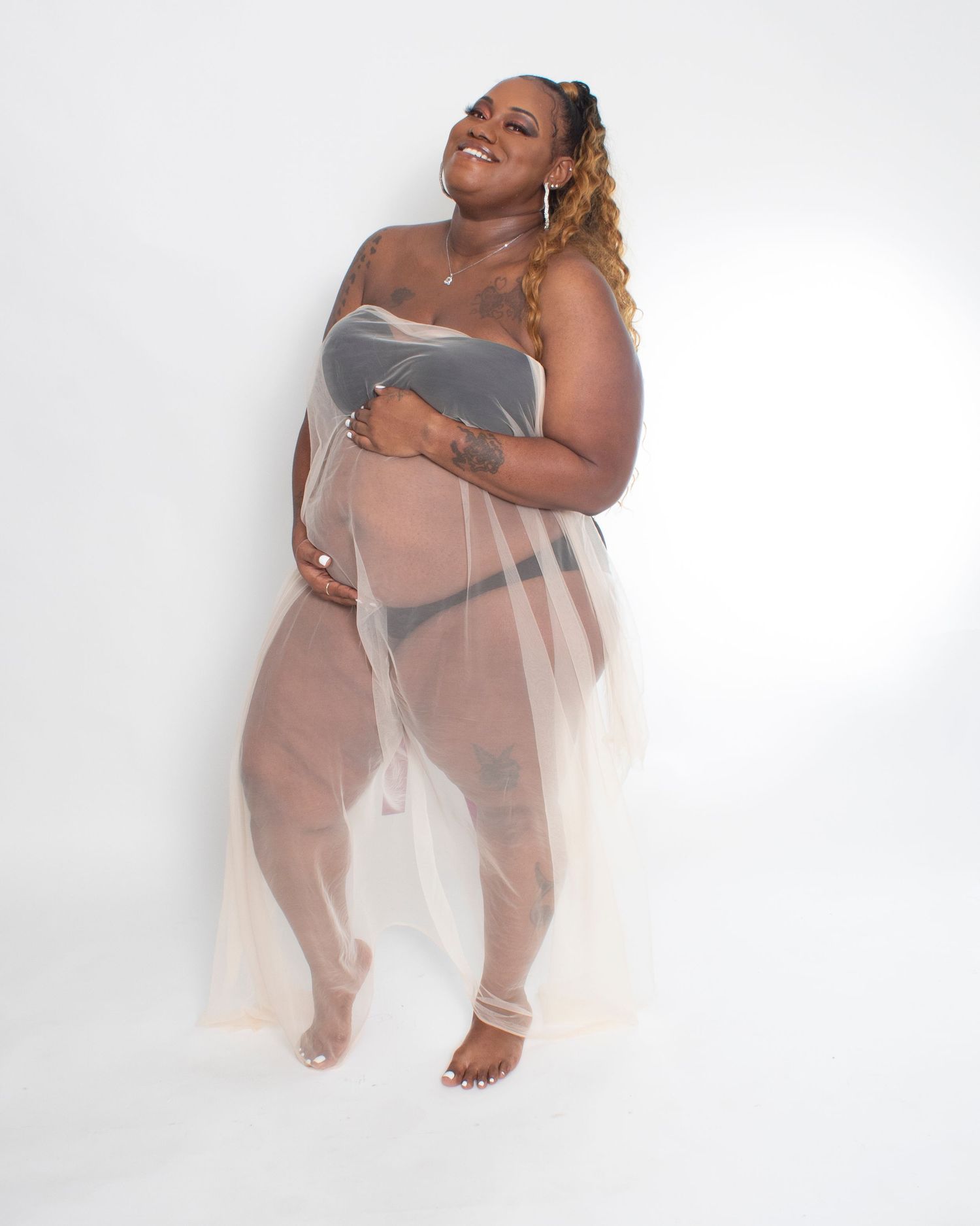 Pregnant Black woman in gray strapless top with sheer tulle skirt laughing while cradling baby bump.